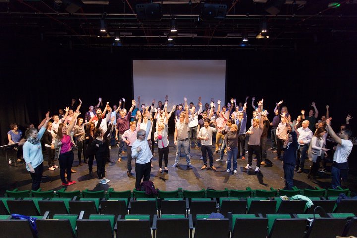 © J Tucker A large group of RADA Business course participants take part in a singing workshop in a theatre.
