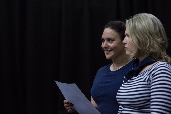Two women stand in front of an audience; one holds a piece of paper.