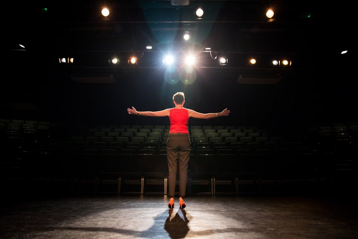 A RADA Business participant stands centre stage in a theatre auditorium. Her arms are outstretched and she is wearing a red top and black trousers.
