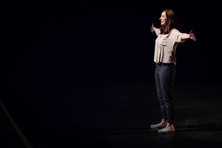 A RADA Business delegate stands on a stage, smiling. Her arms are outstretched.