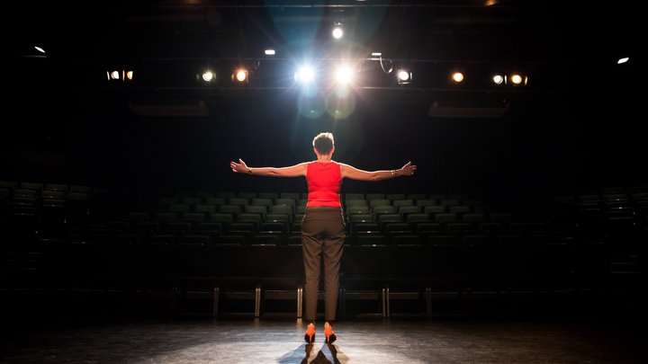 A woman stands on a stage with her back to the camera, illuminated by stage lights, her arms spread wide