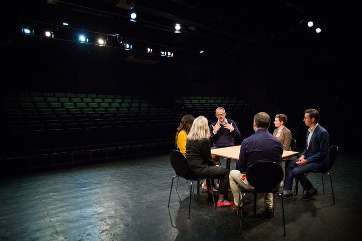 A group of participants sit in a large theatre auditorium around a table. The space is lit with stage lights, hanging from the ceiling.
