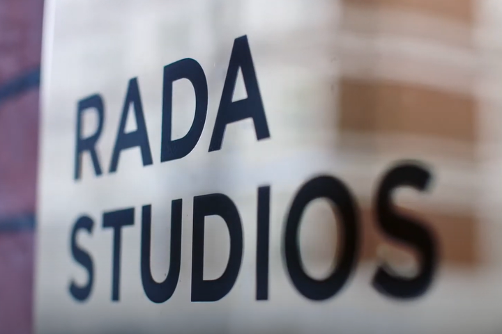 A gold sign on a brick wall with text. The text reads: RADA Studios in black writing.
