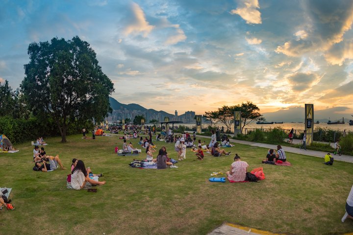 People are sitting on a grass bank that overlooks West Kowloon Cultural District.
