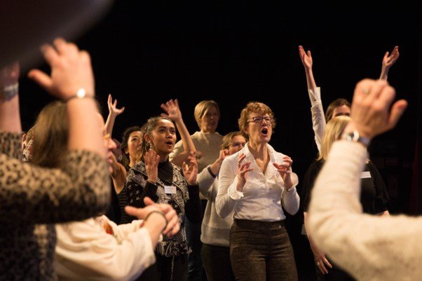 Members of the RADA Business Women's Network take part in a singing workshop in a theatre. They stand in a semicircle.