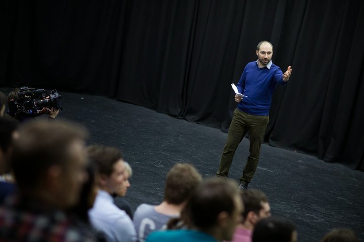 © J Turner A RADA Business tutor presents to a group of business people in a theatre space. His left arm is outstretched toward the audience.