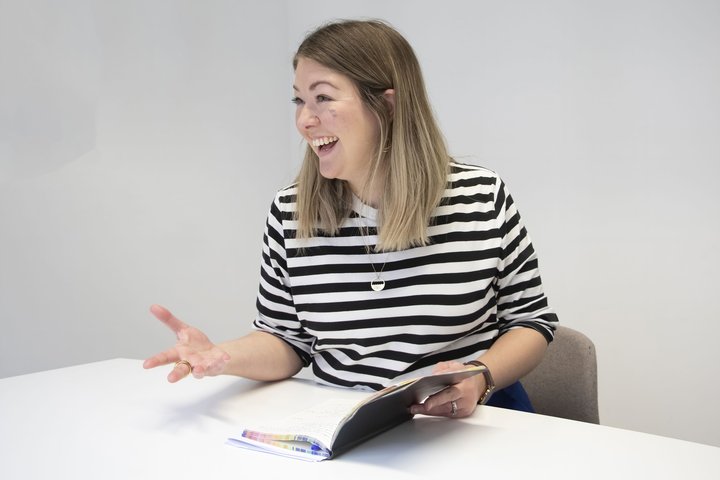 A women with shoulder length blond hair and a stripy t short, sits at a table and gestures.